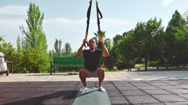 Spor giyimli bir adam direniş bantlarını kullanarak çömelme hareketleri yapıyor, fitness 'e bağlılığını ve açık hava yaşam tarzını gösteriyor..