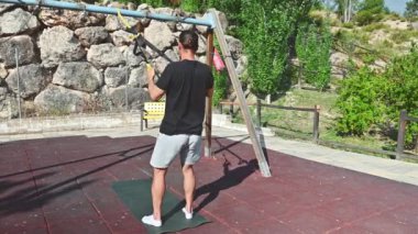 Siyah gömlekli ve gri şortlu bir adam parkta salıncağa bağlı egzersiz direnci bantları kullanarak çömelme hareketleri yapıyor. Bu fitness videosu odaklanmış bir sporcunun eşsiz bir şekilde spor yaptığını gösteriyor..