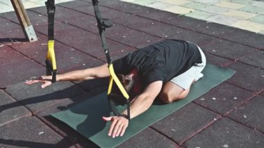 Bir adam, TRX süspansiyon antrenörünü kullanarak kollarını ve sırtını uzatıyor. Bir paspasın üzerinde diz çöküyor ve kollarıyla uzanıyor. Formda ve sağlıklı olduğunu gösteriyor..