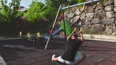 Spor kıyafetleri giymiş bir adam güneşli bir günde açık havada egzersiz platformunda bir dizi spor yapıyor. Direniş eğitimi için kayışlar kullanıyor..