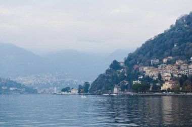 Como Gölü manzarası çok güzel. Tepe kenarındaki evleri ve arka planda sisli dağları var. Sakin sular ve pitoresk manzaralar dingin bir atmosfer yaratır..