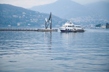 Feribot, sakin bir göl kenarındaki modern bir heykelin yanında demirlemiş. Etrafı güzel bir yamaçla çevrili. Sakin su, sisli atmosfer ve tepe tepe arkaplan sakin bir sahne oluşturur.