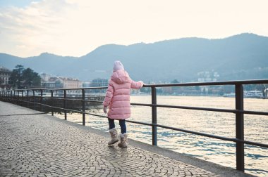 Pembe kışlık paltolu genç kız gölün kenarında duruyor, arkasında dağlar var. Sakin suları ve uzak dağları olan huzurlu kış manzarası.