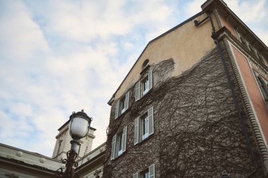 Mavi gökyüzünün altında dağınık bulutlu beyaz panjurları olan sarmaşık kaplı tarihi bir bina. Klasik mimari ve sokak lambası eski bir Avrupa şehir atmosferini ortaya koyuyor.