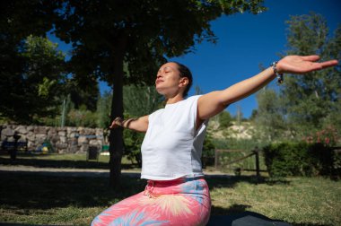 Kadın açık havada kollarını açarak yoga yapıyor. Ağaçlar ve yeşilliklerle güneşli bir gün.