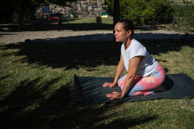 Parkta güneşli bir günde yoga yapan bir kadın sağlığını, rahatlığını ve farkındalığını gösterir..