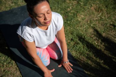 Paspasın üzerinde yoga yapan bir kadın, gün ışığında huzur ve rahatlığın tadını çıkarıyor. Beyaz bir bluz ve renkli taytlar giyiyor..