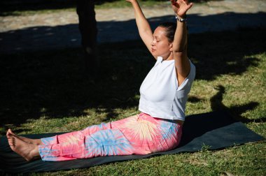 Dışarıda yoga yapan bir kadın, sabahın köründe elleri havada bir paspasın üzerinde oturuyor. Odaklanmış ve sakin görünüyor..