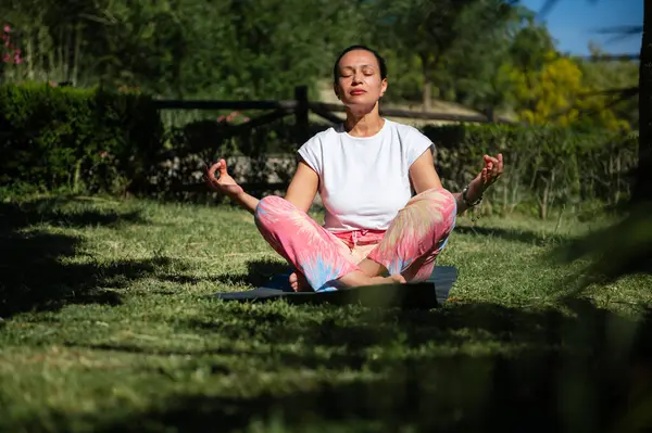 Renkli pantolonlu bir kadın, açık mavi bir gökyüzünün altında, açık bir bahçede, bir paspasın üzerinde bacak bacak üstüne atmış, meditasyon yapıyor..