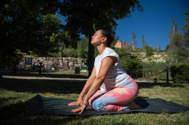 Huzur parkında yoga yapan bir kadın. Bir ağacın gölgesinde yoga minderinde renkli taytlar giyiyor ve açık havanın tadını çıkarıyor..