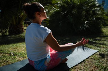 Dışarıdaki yoga minderinde oturmuş, sakin bir bahçe ortamında meditasyon yapan bir kadın. Günlük atletizm kıyafetleri içinde ve huzurlu bir ortamın tadını çıkarıyor..