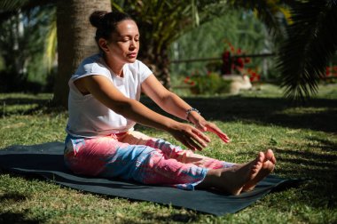 Renkli pantolonlu bir kadın yoga yapıyor. Açık hava ortamında paspasın üzerinde geriniyor. Etrafı yeşillikle çevrili..