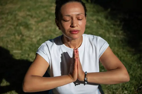 Stock image A woman in a white shirt practices yoga and meditation outdoors with her eyes closed, hands in prayer position, enjoying a peaceful moment.