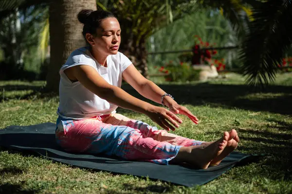 stock image Woman in colorful pants practicing yoga stretches on a mat in a serene outdoor setting, surrounded by greenery.