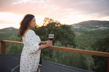 Bir bardak kırmızı şarapla balkonda duran kadın manzaraya ve günbatımına hayran. Sakin ve huzurlu bir atmosfer..