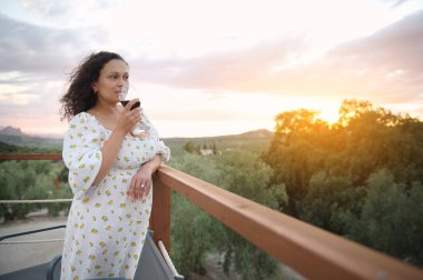 Beyaz elbiseli bir kadın balkonda kırmızı şarap içiyor gün batımını ve doğayı manzaralı, rahat ve huzurlu hissediyor..
