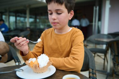 Sarı tişörtlü bir çocuk, açık hava kafesinde waffle külahında dondurma yiyip, tatlı bir zevk anı yaratıyor..