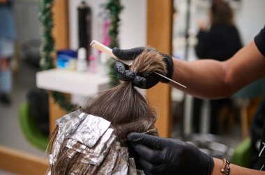 Eldiven giyen bir kuaför, müşterinin saçına folyo ve modern bir güzellik salonunda boya kullanarak röfle uygular. Profesyonel saç boyama hizmetleri.