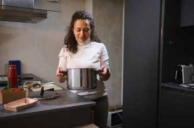 Kadın modern mutfağında yemek hazırlıyor. Elinde paslanmaz çelik bir tencere var. Taze malzemeler ve mutfak gereçleri tezgahta, rahat ve davetkar bir atmosfer yaratıyorlar..