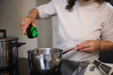 Close-up of a woman pouring oil into a pot while cooking in a modern kitchen. Concept of home cooking and healthy meal preparation. clipart