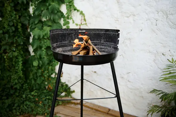 stock image A black round fire pit standing against a green leafy background with burning wood, perfect for outdoor gatherings and cozy evenings.