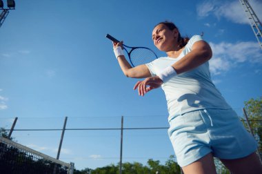 Açık hava kortunda tenis oynayan kararlı bir kadın. Arka planda açık mavi bir gökyüzü ile salıncağın ortasında..