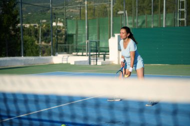 Dışarıda tenis oynayan bir kadın, servis yapmaya hazırlanıyor. Spor kıyafetleri giyerek, o oyuna odaklandı. Güneşli hava ve yeşil çevre enerjiyi artırır..