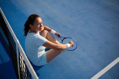 Beyazlar içinde bir tenis oyuncusu mavi bir tenis kortunda oturuyor, dinleniyor ve elinde bir raket, odaklanmış görünüyor..
