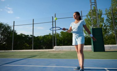 Spor kıyafetli bir kadın açık mavi gökyüzü olan bir kortta tenis oynuyor. Odaklandı ve topa vurmaya hazır..