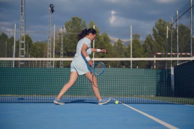 Spor giyim sektöründe tenis oynayan genç bir bayan, spor yeteneklerini sergiliyor. Sahne onun orta adımlarını yakalar ve oyuna odaklanır, fitness ve aktif yaşam tarzı temalarına katkıda bulunur..