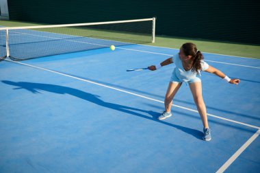 Açık hava mavi tenis kortunda tenis topuna vuran hareketli genç bir kadın çeviklik ve kararlılık gösteriyor..