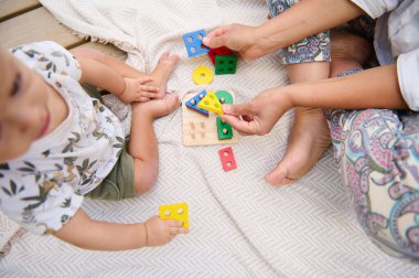 Bir anne ve çocuk, renkli geometrik şekillerle battaniyenin üzerinde oynayarak eğlenceli ve eğitici bir aktiviteye girişiyor. Öğrenme, bağlanma ve erken çocukluk gelişimiyle ilgili kavramlar için mükemmel.