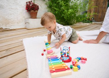Bebek, tahta bir güvertede, battaniyenin üzerinde canlı ahşap oyuncaklarla oynuyor. Yakınındaki ebeveyn destek öneriyor.