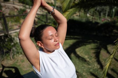 Bir kadın yoga yaparken gözleri kapalı, huzurlu bir şekilde meditasyon yapar. Doğa ortamında, rahatlama ve farkındalığı somutlaştırır..