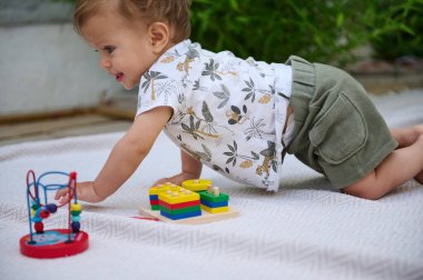 Güneşli bir günde açık havada renkli oyuncaklarla oynarken beyaz oyun hasırında sürünen desenli tişörtlü ve yeşil şortlu şirin bir bebek..