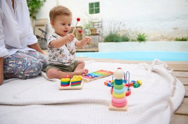 Havuz kenarındaki battaniyenin üzerinde renkli oyuncaklarla oynayan sevimli bir bebek. Ebeveyn bebeğin yanında oturuyor. Çocukluk gelişimi ve bağ kurmak için ideal ortam..