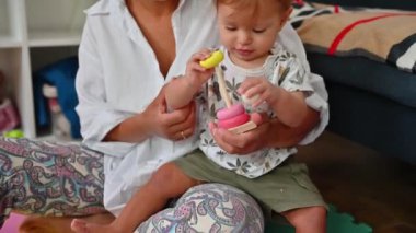 Sevimli erkek bebek renkli yuvarlak tahta piramitlerle oynuyor, onları tadıyor, annesinin ellerinde oturuyor. Yeni yürümeye başlayan çocuk dokunma ve tatma ile çevre öğreniyor. Düşünce ve motor beceriler geliştiriyor