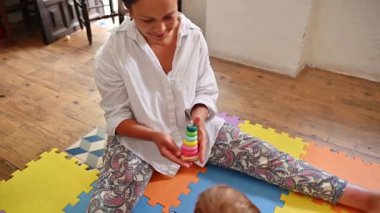 Genç anne gülümsüyor, elinde ahşap bir piramit tutuyor, bebeğini daireler çizmeye teşvik ediyor, renkli bir yapboz halısında birlikte oturuyorlar. Doğum izni ve çocuk gelişimi konsepti