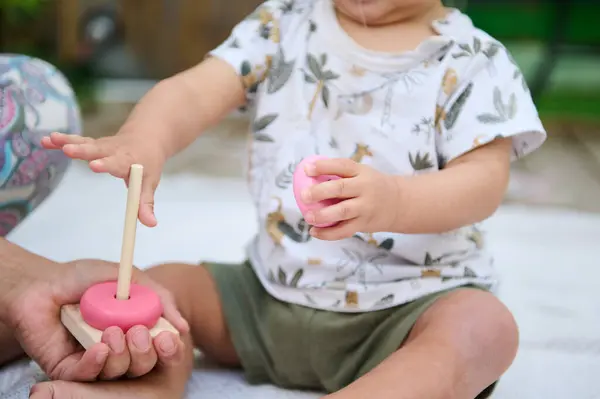 stock image Close-up with focus on hands of a baby boy stacking round circles of educational developmental wooden pyramid, playing with his mother supervising him, sitting together on the linen blanket outdoors