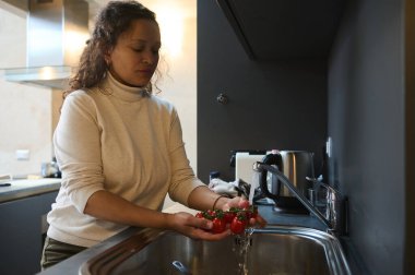 Mutfak lavabosunun üzerinde kiraz domatesleri ve diğer sebzeleri yıkayan çok ırklı, sağlıklı genç bir kadın kirlenme riskini ortadan kaldırmak için suyla temizlik yapıyor.