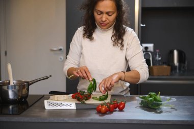 Elinde taze fesleğen yaprağı tutan çok ırklı bir kadın çiğ vegan salatası hazırlıyor. Mutfak tezgahında bir demet sağlıklı vişneli domates ve bir parça parmesan peyniri görebiliyoruz. - İnsanlar. Yiyecekler. Yaşam biçimi