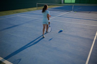 Dış sahada tenis oynayan bir kadının eğitim makinesiyle arkadan görünüşü. Fitness, eğitim ve açık hava spor faaliyetlerine vurgu yapılıyor.