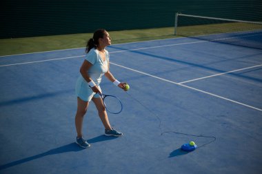 Bir kadın güneşli bir günde açık mavi tenis kortunda tenis yeteneklerini geliştiriyor. Raketiyle tenis topuna vurmaya hazırlanıyor..