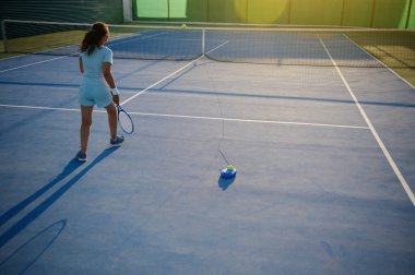 Genç bir kız tenis antrenörü kullanarak açık hava kortunda tenis oynuyor. Sıcak güneş ışığı ilham verici ve motive edici bir atmosfer yaratır..