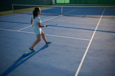 Açık hava mavi tenis kortunda antrenman yapan genç bir bayan tenisçi. Yeteneklerini ve tekniğini geliştirmeye odaklanmış..