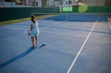 Açık havada tenis kortunda raket ve topla yeteneklerini sergileyen bir bayan tenisçi. Mahkeme boş ve sahne alacakaranlıkta çekiliyor..