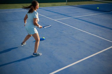 Gündüz vakti mavi kortta tenis oynayan bir kadın topa vurmaya odaklanıyor. Atletik ve enerjik açık hava aktivitesi.