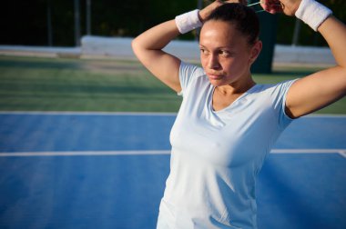 Odaklanmış bayan tenisçi güneşin altındaki maça hazırlanırken saçını mavi kortta bağlıyor..