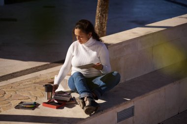 Dışarıdaki taş bir bankta oturan bir kadın, etrafında malzemelerle tablet çiziyor, güneşli bir günün tadını çıkarıyor..