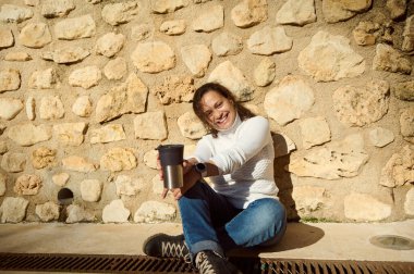 Kırsal taştan bir duvara yaslanmış mutlu bir kadın elinde kahve fincanı, parlak bir gülümsemeyle açık havada güneşli bir günün tadını çıkarıyor..